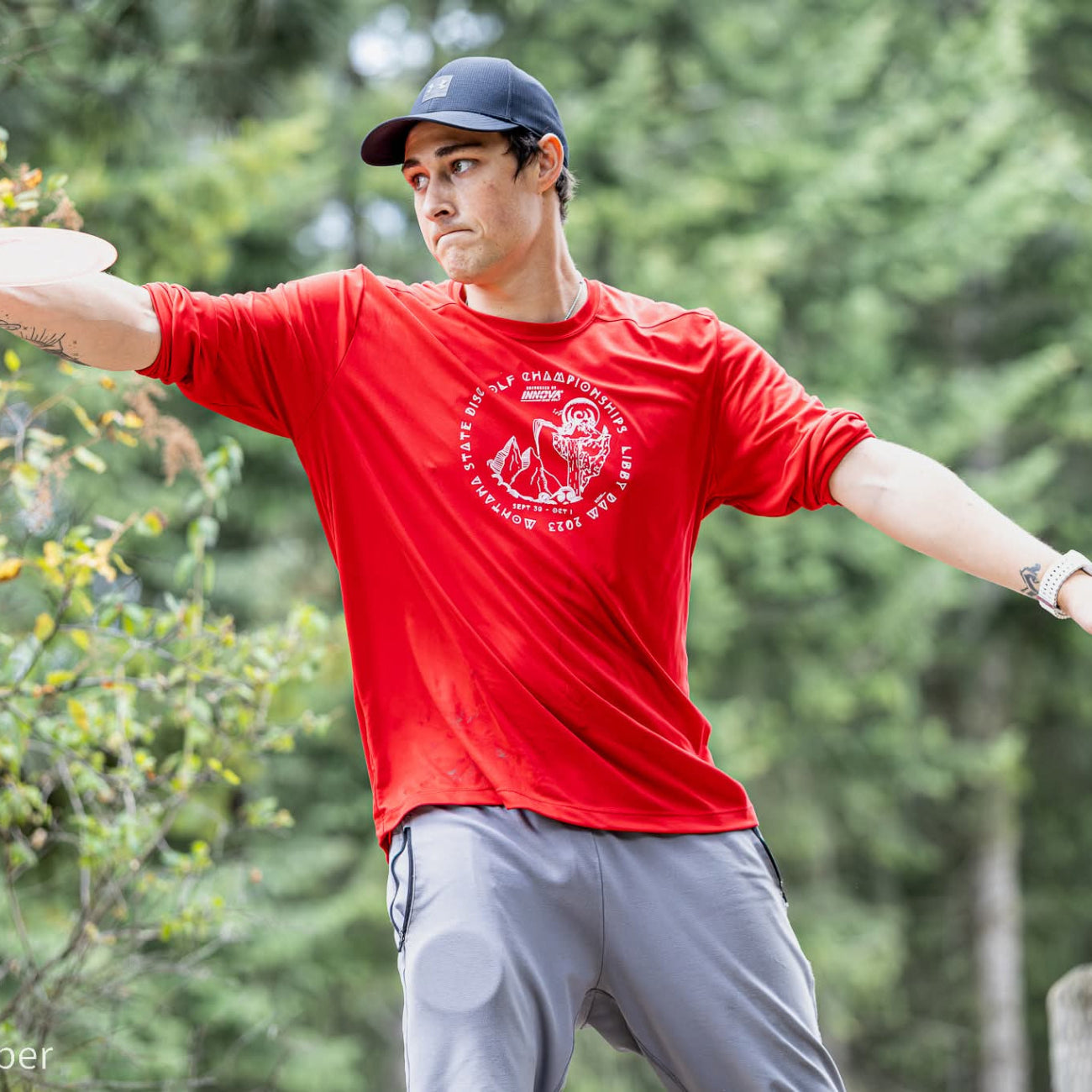 Man in a red shirt and gray pants throwing a frisbee outdoors with trees in the background