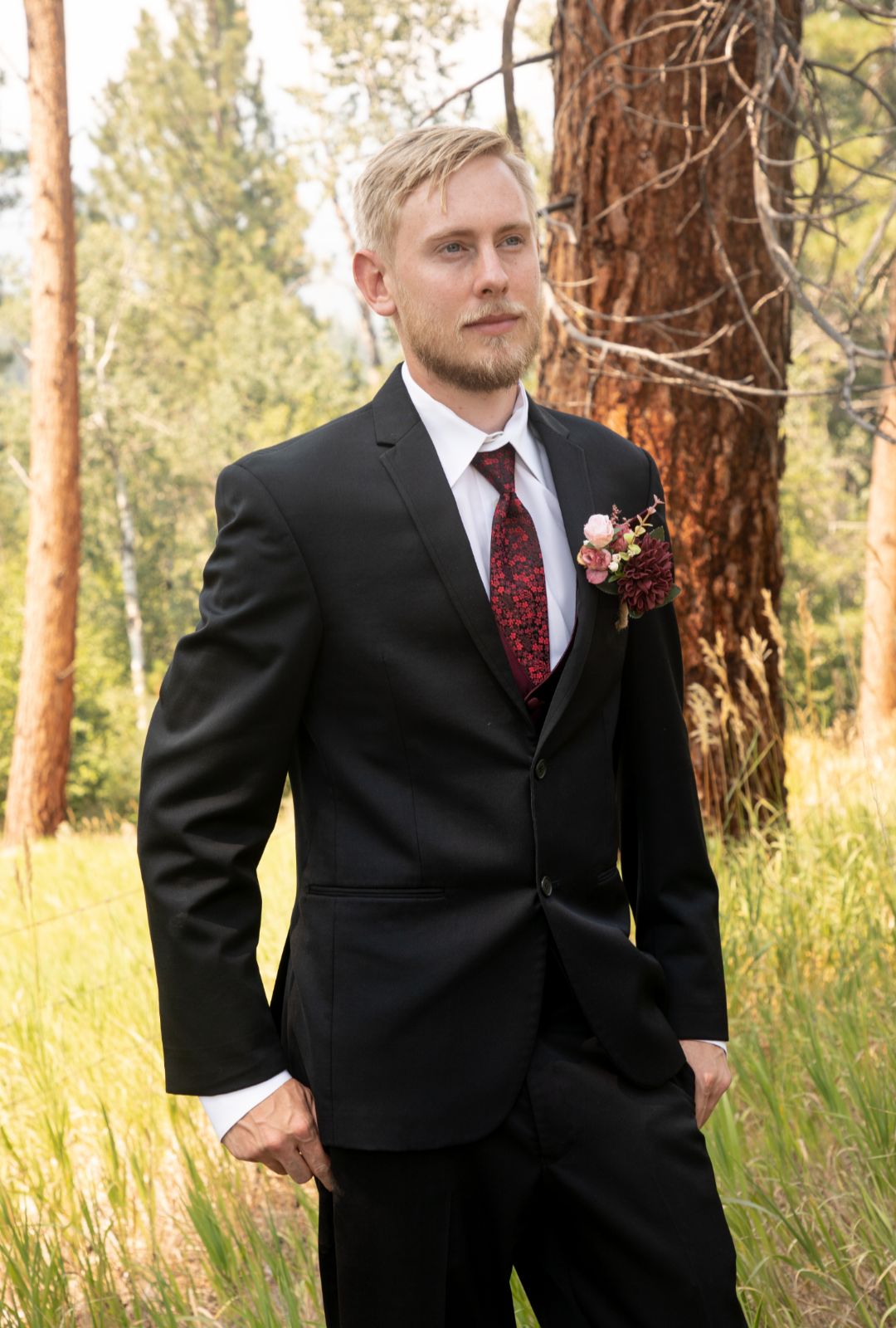 Man in a black suit with a red tie and boutonniere standing in a forest.