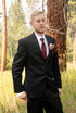 Man in a black suit with a red tie and boutonniere standing in a forest.
